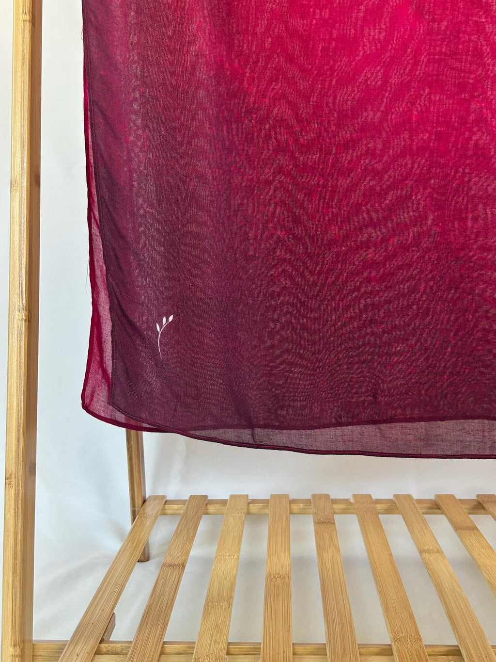 A sheer maroon ombre scarf with a small white leaf embroidery hangs gracefully from a bamboo clothing rack.