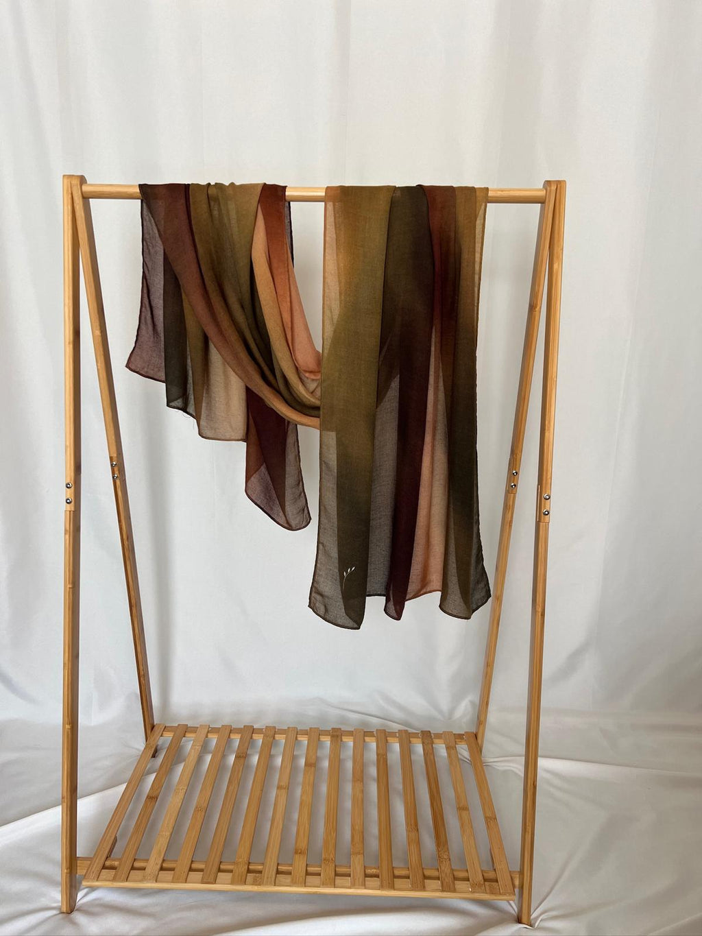 Two lightweight sheer scarves in earthy shades of olive, brown, and rust drape elegantly over a wooden bamboo rack against a white backdrop.