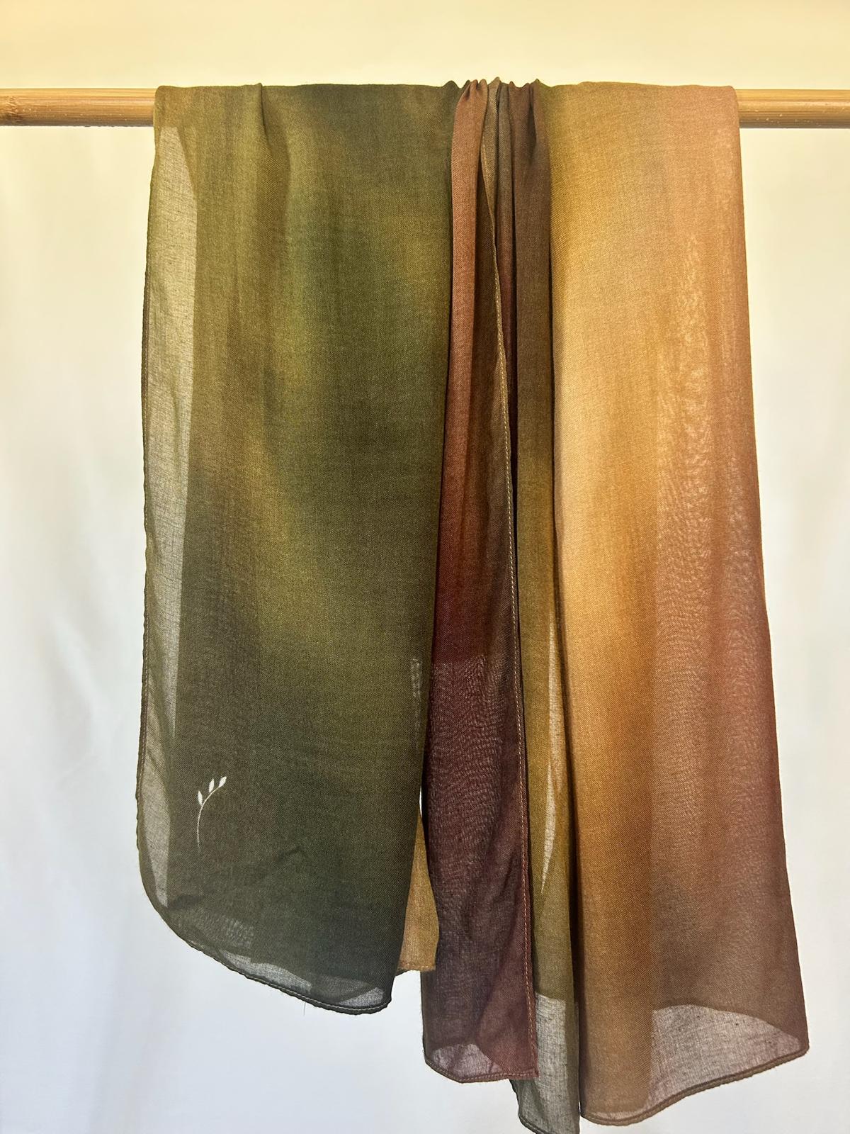 Lightweight, earth-toned ombré scarves in shades of olive green, amber, and brown hang gracefully from a wooden rod.