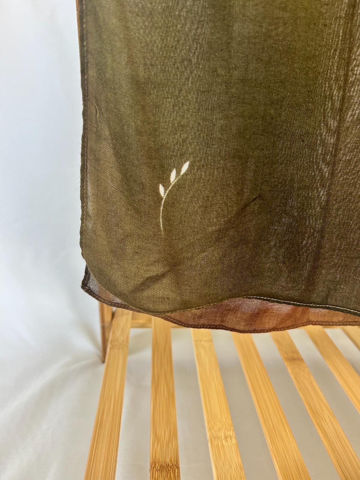 A sheer olive green scarf featuring a delicate white leaf motif hangs over a light wood bamboo slatted rack.