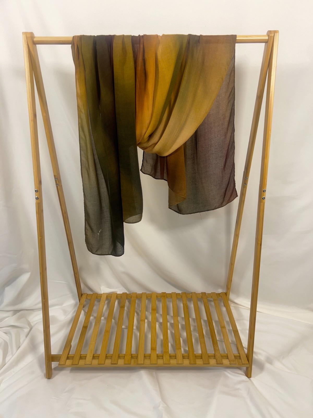 A lightweight ombre scarf in warm shades of gold, olive green, and dark brown drapes over a minimalist wooden garment rack.