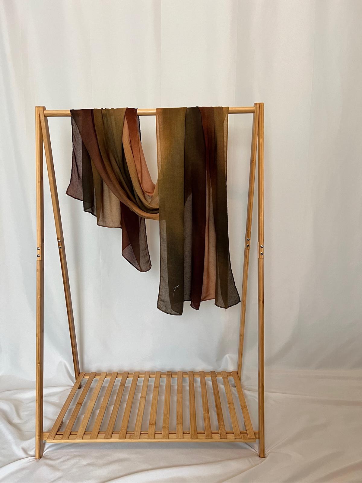 A sheer, earthy-toned scarf in shades of brown and olive green drapes elegantly over a light wood garment rack against a clean white backdrop.