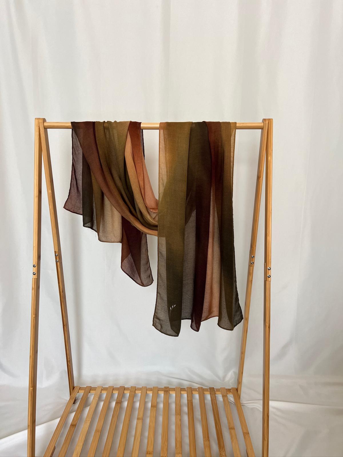 Two lightweight, sheer scarves in earthy autumn tones hang elegantly over a wooden garment rack against a soft white background.