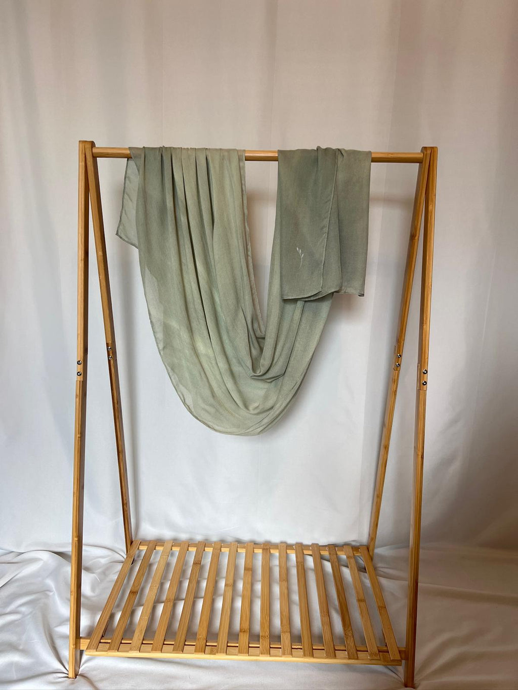 A sage green hand-dyed scarf drapes gracefully over a minimal bamboo clothing rack against a soft white background.