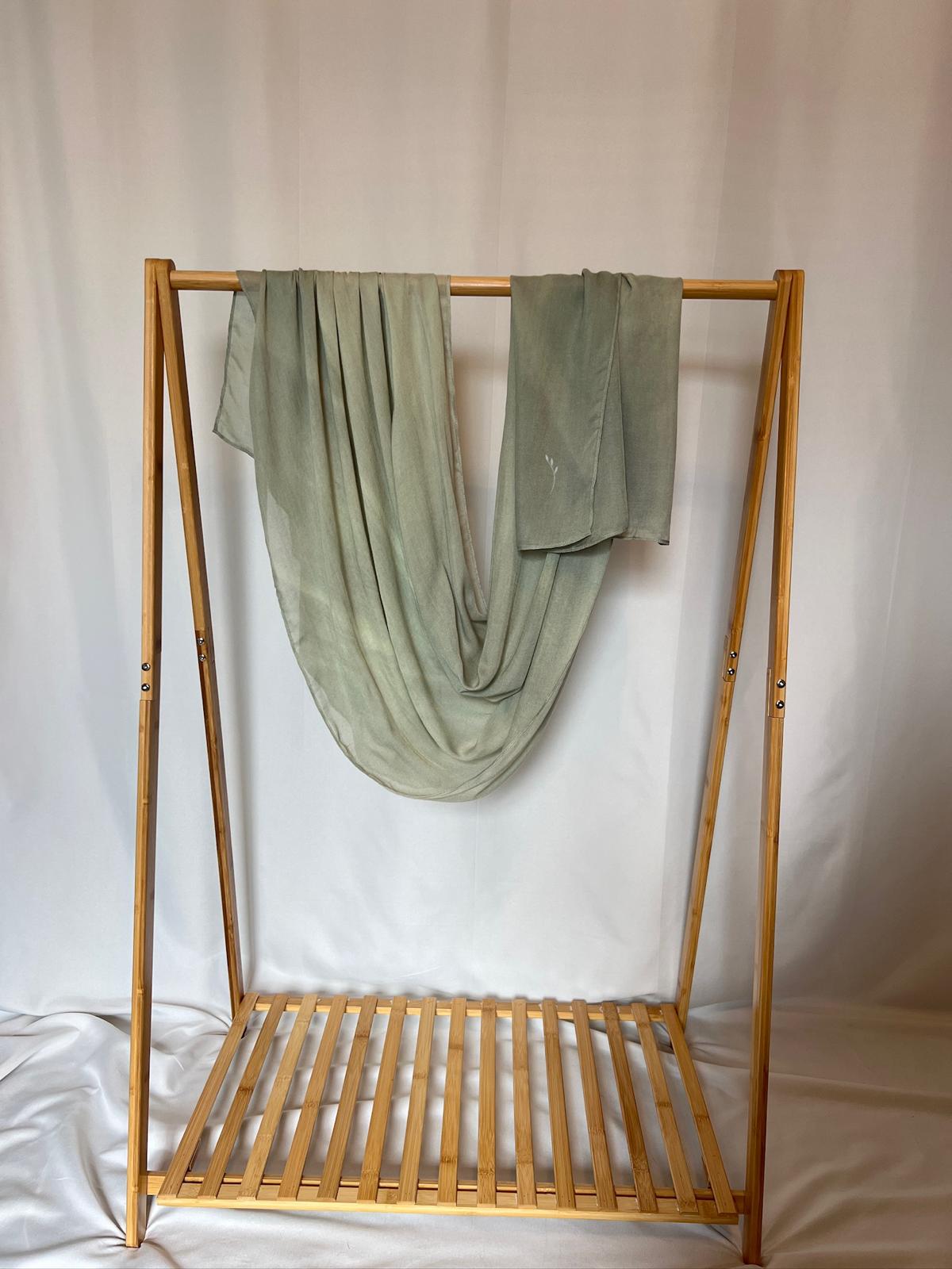 A sage green hand-dyed scarf drapes gracefully over a minimal bamboo clothing rack against a soft white background.