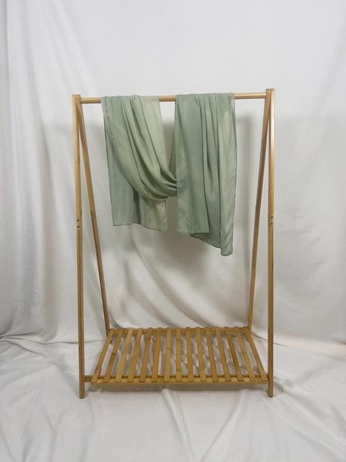 A light sage green sheer scarf hangs gracefully from a minimalist wooden garment rack with a slatted storage shelf.