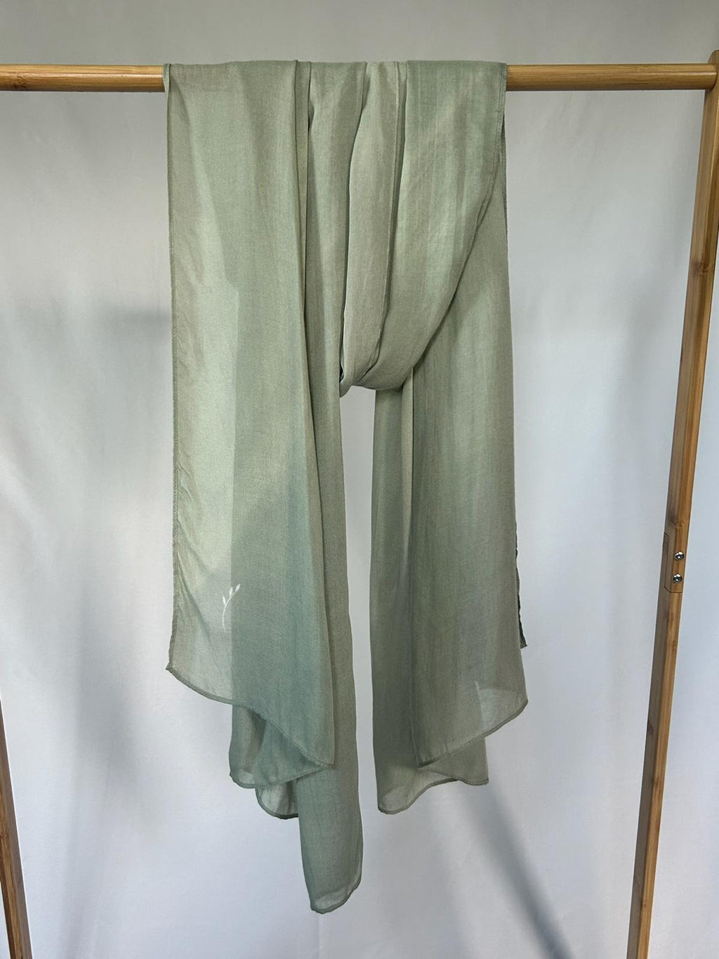 A lightweight sage green semi-sheer scarf with a small white embroidered leaf hangs from a bamboo clothing rack against a plain white background.