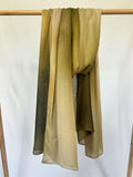 A lightweight, hand-dyed olive green to tan ombre scarf drapes over a wooden rod against a white background.