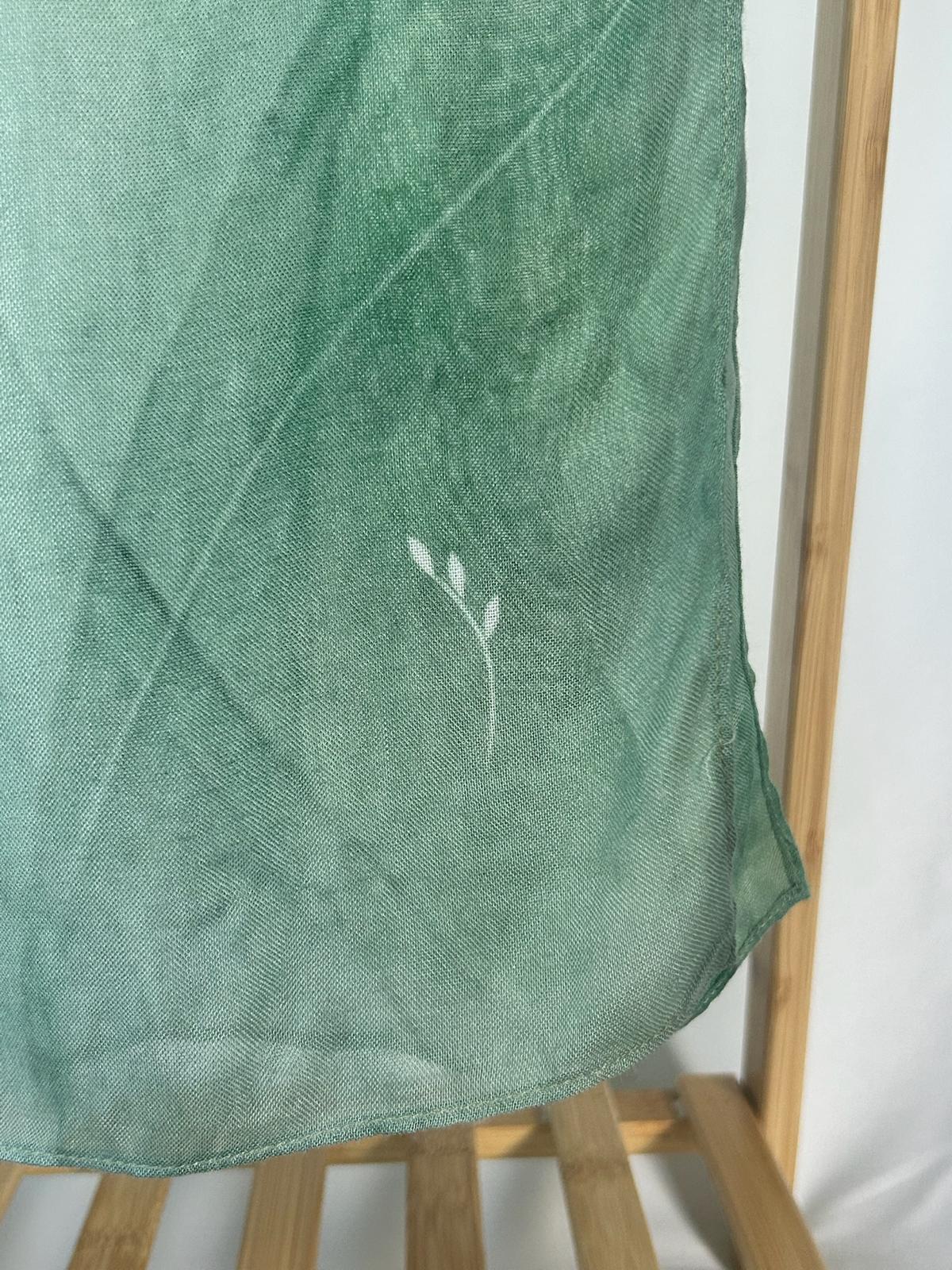A delicate white leaf embroidery accents a sheer sage green fabric hanging from a wooden rack.