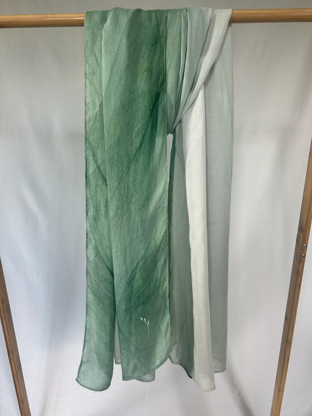 A lightweight sage green ombre scarf with a delicate white leaf detail hangs elegantly on a wooden garment rack.