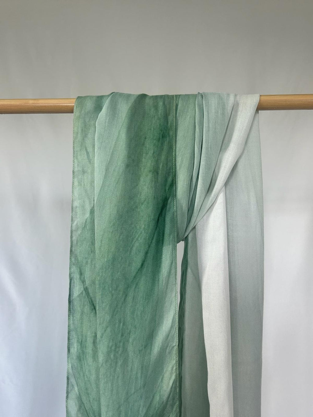 A lightweight green ombre silk scarf drapes elegantly over a wooden rod against a clean white background.