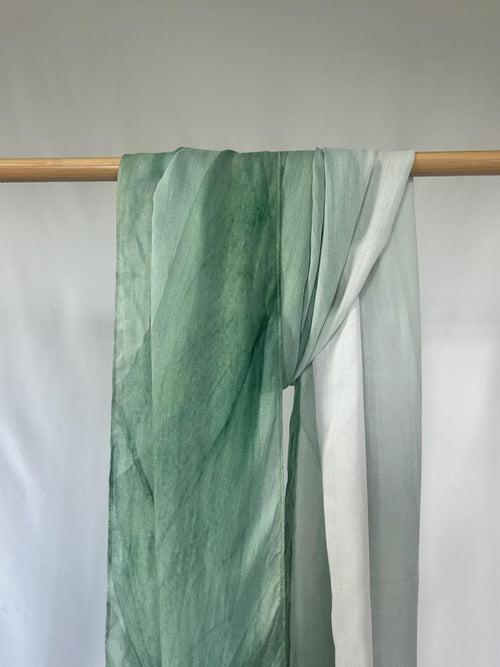A lightweight green ombre silk scarf drapes elegantly over a wooden rod against a clean white background.