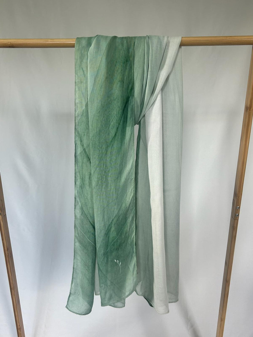 A sheer green silk scarf with a subtle gradient pattern and a delicate leaf emblem hangs from a wooden display rack.