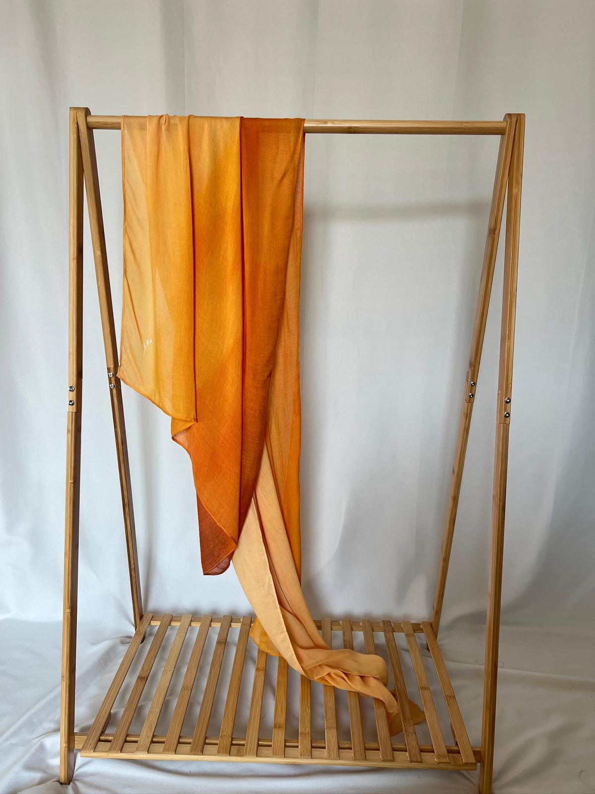A lightweight orange ombré scarf drapes gracefully over a minimalist wooden garment rack against a white background.