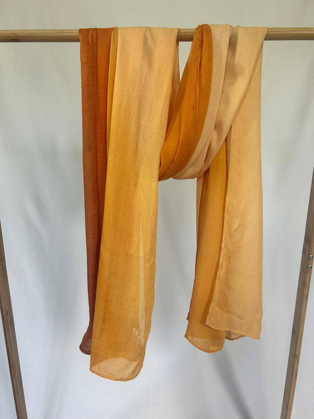 A lightweight silk scarf with an amber and orange gradient drapes gracefully over a wooden garment rack.