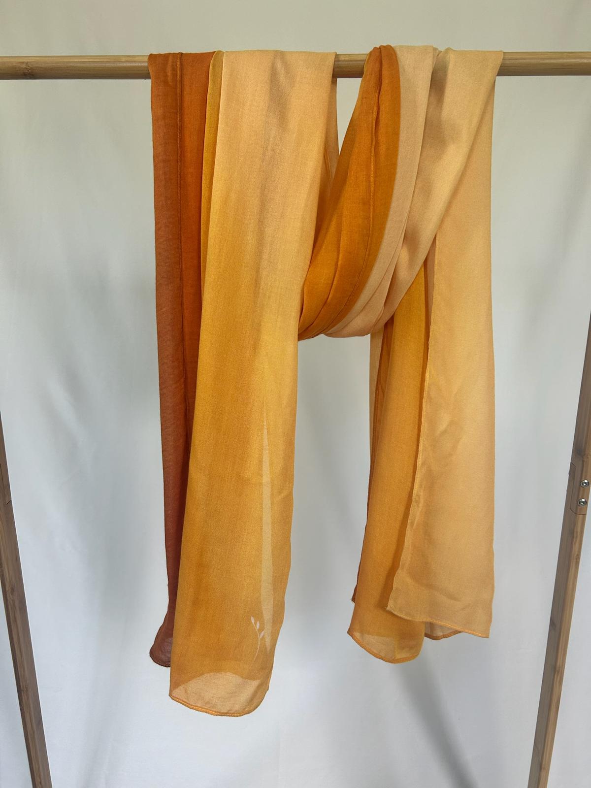 A lightweight silk scarf with an amber and orange gradient drapes gracefully over a wooden garment rack.