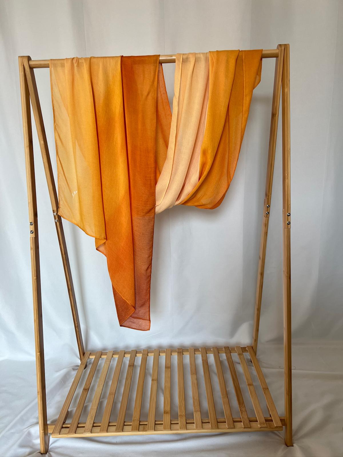 A vibrant orange and yellow ombre scarf drapes elegantly over a minimalist bamboo clothing rack.